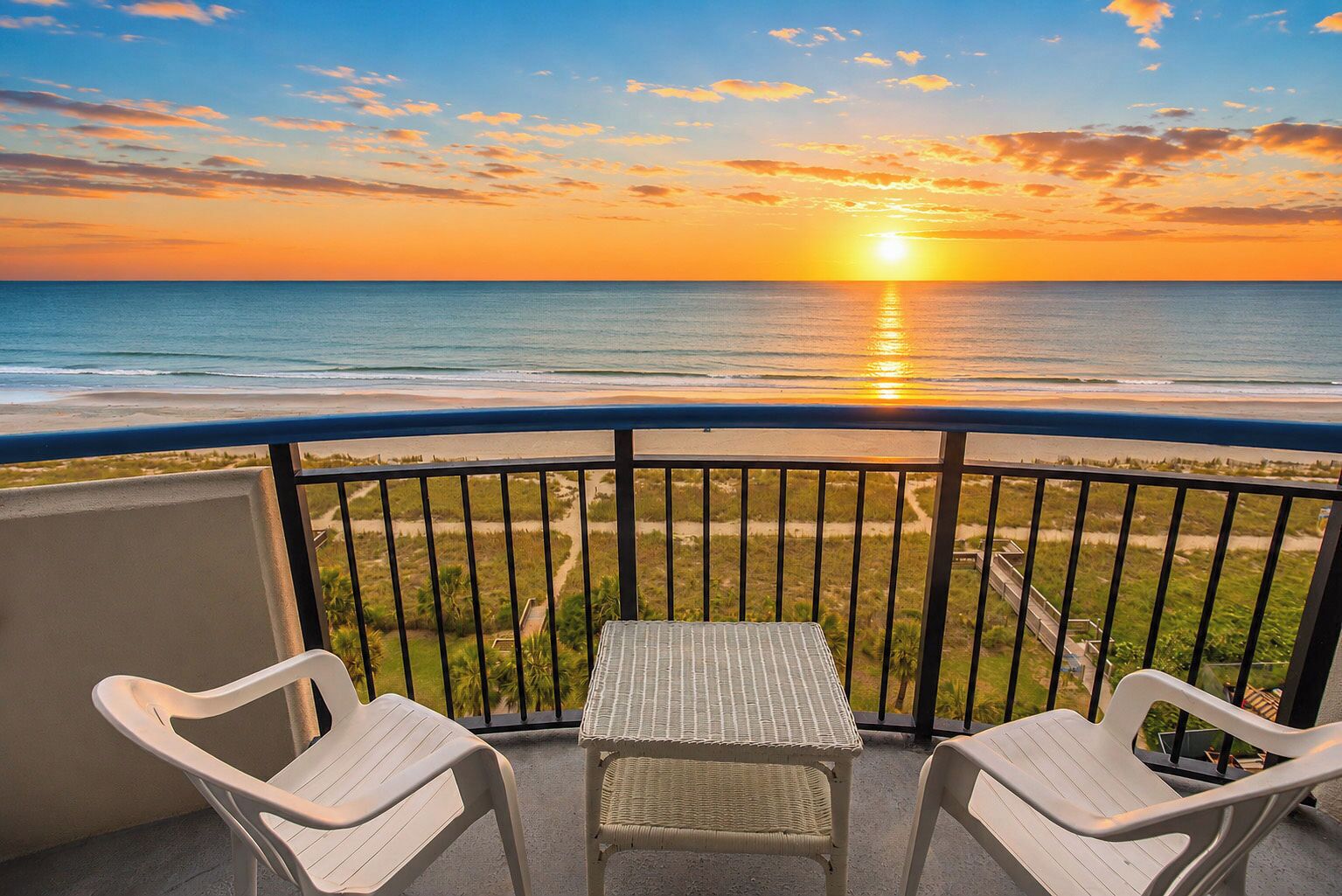 A balcony with two chairs and a table, overlooking a beach and ocean during sunset.