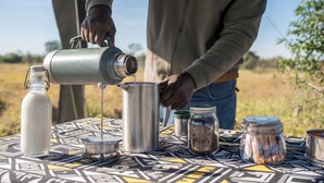A person pouring liquid from a metal container into a stainless steel pot on a patterned table outdoors. - Satara Wild Camp- Greater Kruger (Mpumalanga)