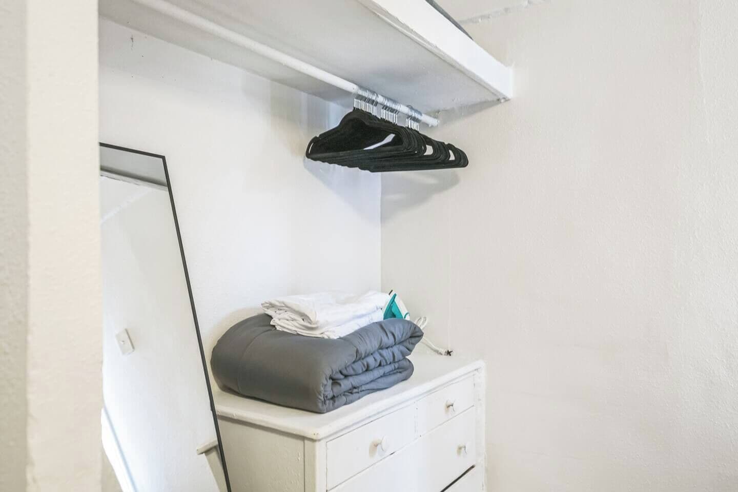 A white dresser with a grey cushion and folded clothes, a mirror, and a black hanger on a white shelf.