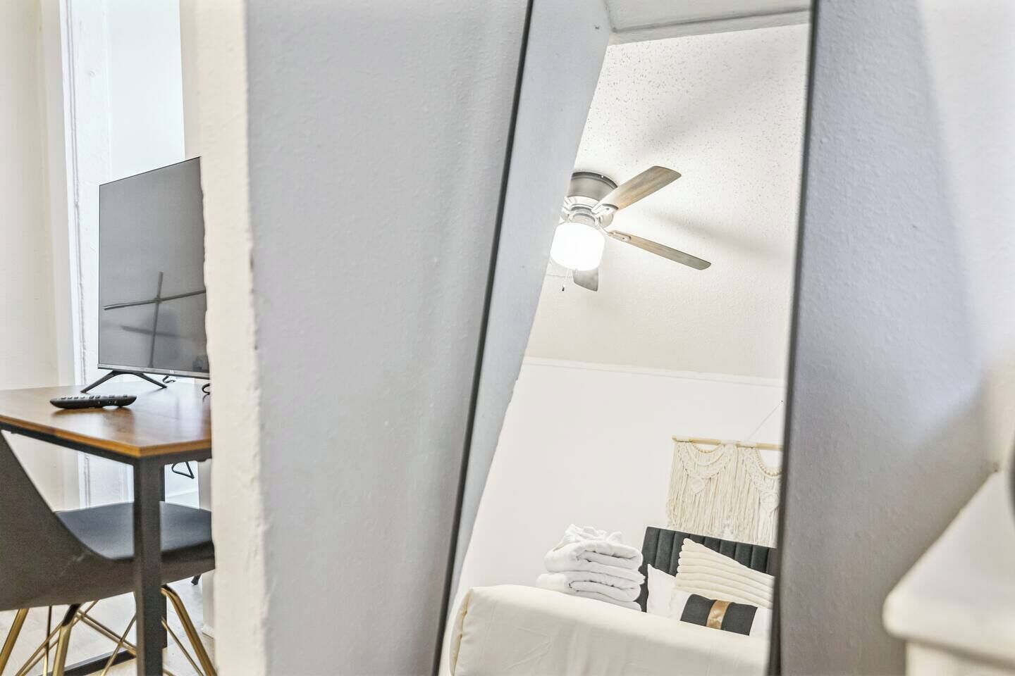 Um quarto com escrivaninha, monitor de computador, ventilador de teto e uma cama com toalhas dobradas.