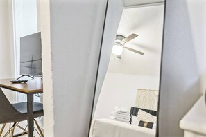 Una stanza con una scrivania, un monitor del computer, una ventola a soffitto e un letto con asciugamani piegati con cura.