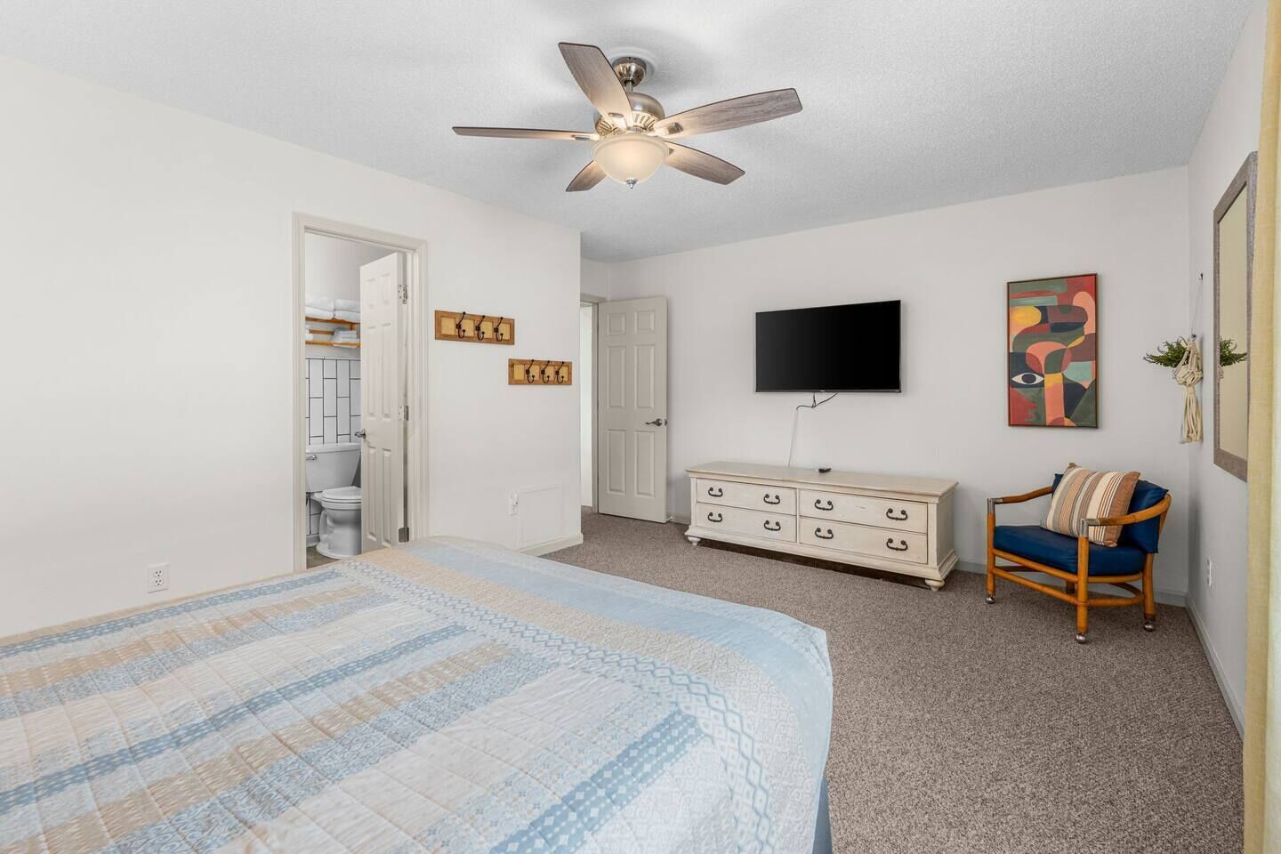 Un dormitorio con cama, cómoda, silla, ventilador de techo, televisor y puerta que da al baño.
