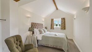A bedroom with a bed, a nightstand, a lamp, a chair, and a window with curtains. - Glebe Cottage - sleeps 4 guests  in 2 bedrooms, single storey property, Tetbury (Near Tetbury)
