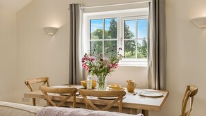 A dining area with a table set for four, a vase with flowers, and a window with curtains. - Glebe Cottage - sleeps 4 guests  in 2 bedrooms, single storey property, Tetbury (Near Tetbury)