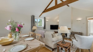 Een woonkamer met een bank, een eettafel gedekt voor vier personen, een aan de muur bevestigde televisie, en een houten balk in het plafond.