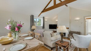 A living room with a sofa, a dining table set for four, a TV mounted on the wall, and a wooden ceiling beam. - Glebe Cottage - sleeps 4 guests  in 2 bedrooms, single storey property, Tetbury (Near Tetbury)