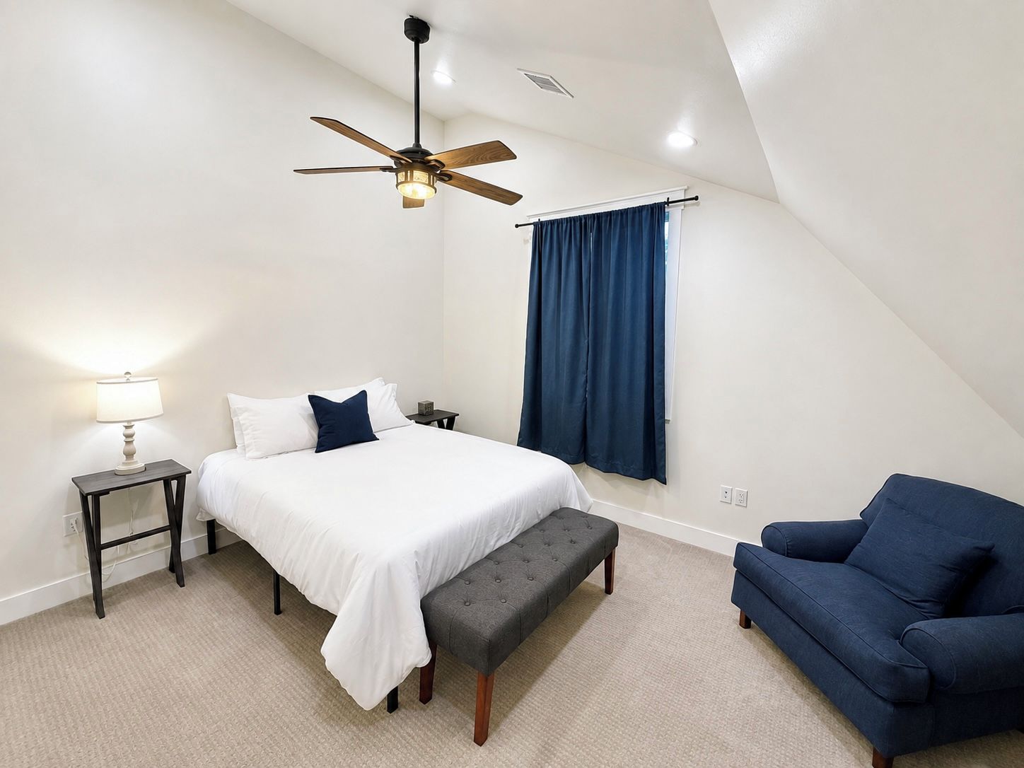 A bedroom with a bed, a bench, a chair, a lamp, a ceiling fan, and a window with curtains.