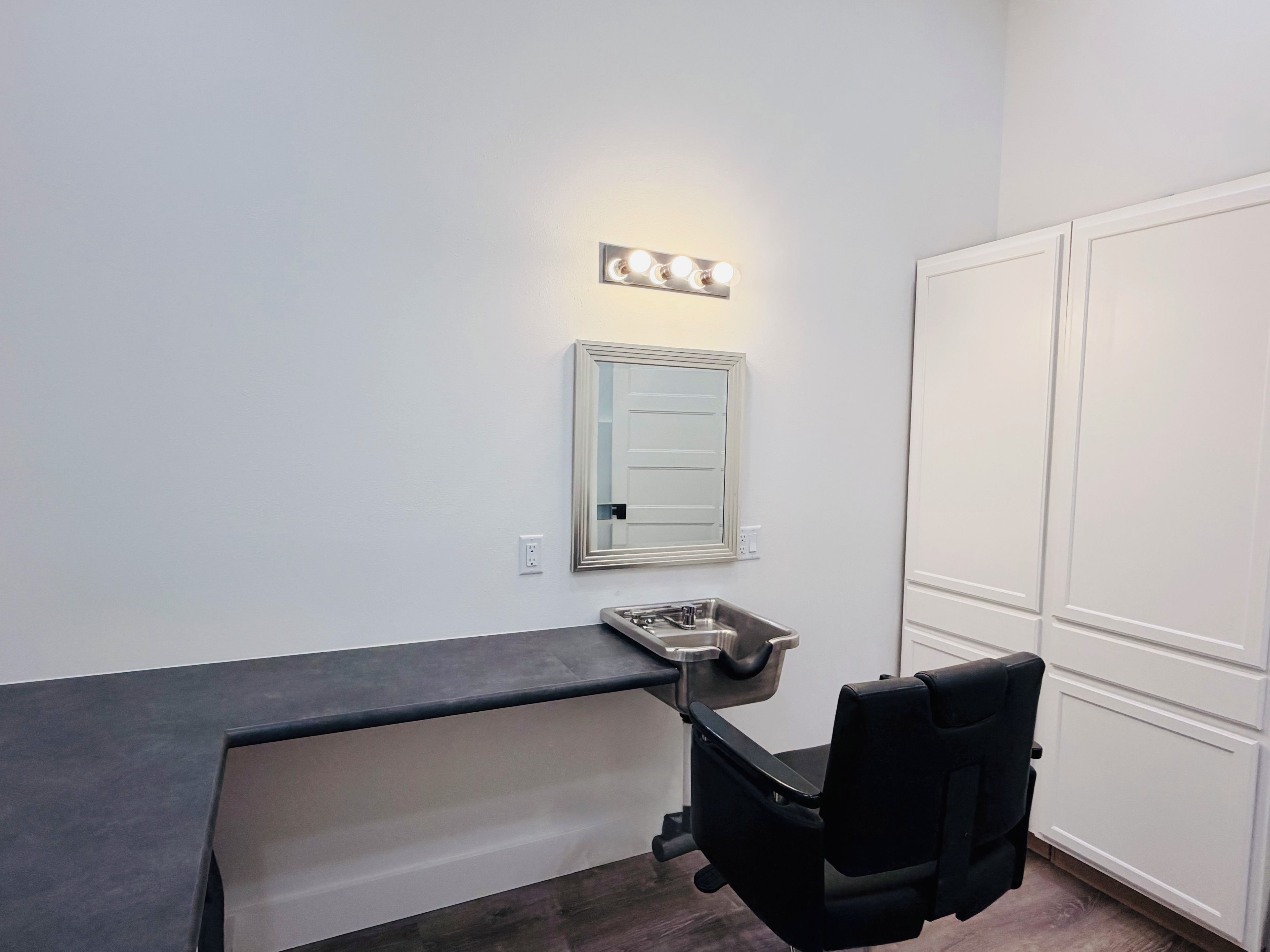 Una habitación con espejo de tocador iluminado, una silla negra y un armario blanco.