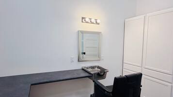 Una habitación con espejo de tocador iluminado, una silla negra y un armario blanco.