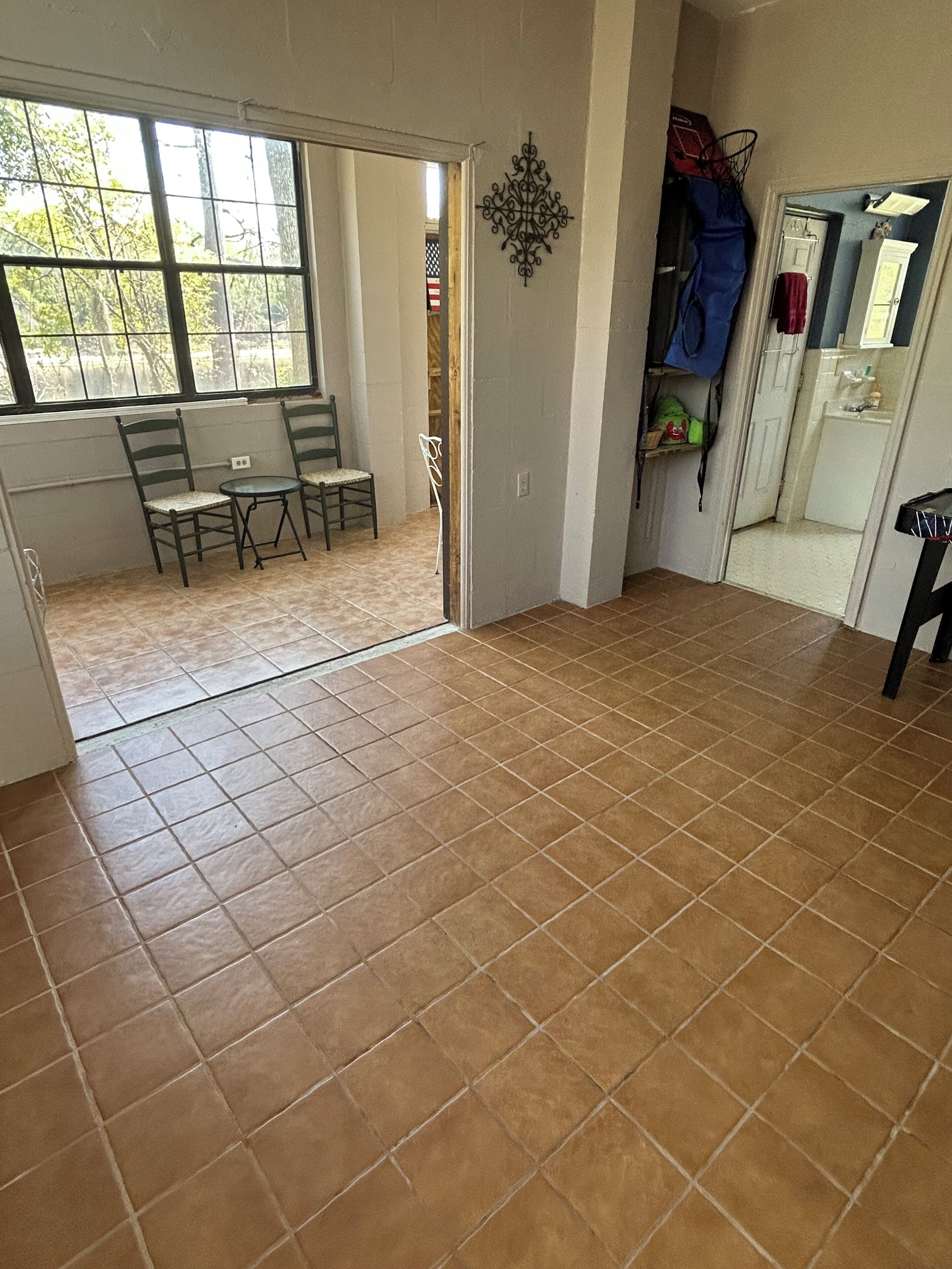 A room with a tiled floor, a dining area with chairs and a table, a door leading to another room, and a wall-mounted decoration.