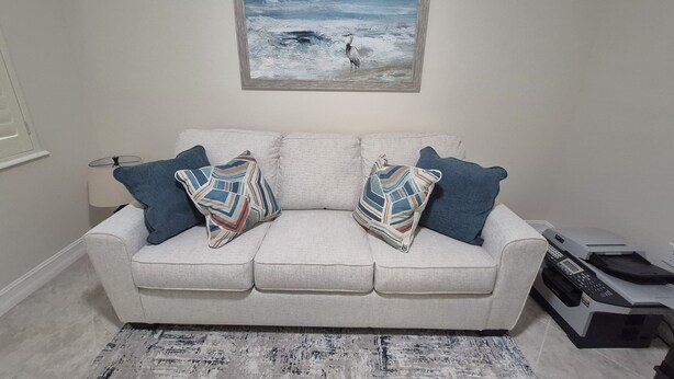 A living room with a beige sofa, patterned pillows, a framed painting of a seascape, a lamp, and a printer on the floor. - 3-bedroom + Den/2 Bath/2 Car Garage (Naples)
