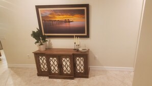A hallway with a wooden console table, a framed painting of a sunset over water, a potted plant, and a candle holder. - 3-bedroom + Den/2 Bath/2 Car Garage (Naples)
