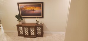 A hallway with a wooden console table, a framed painting of a sunset over water, a potted plant, and a candle holder. - 3-bedroom + Den/2 Bath/2 Car Garage (Naples)