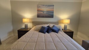 A bed with a beige quilt, two bedside lamps, a nightstand with a tissue box, and a painting of a sunset on the wall. - 3-bedroom + Den/2 Bath/2 Car Garage (Naples)
