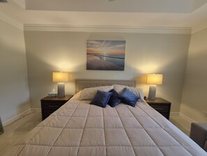 A bed with a beige quilt, two bedside lamps, a nightstand with a tissue box, and a painting of a sunset on the wall. - 3-bedroom + Den/2 Bath/2 Car Garage (Naples)