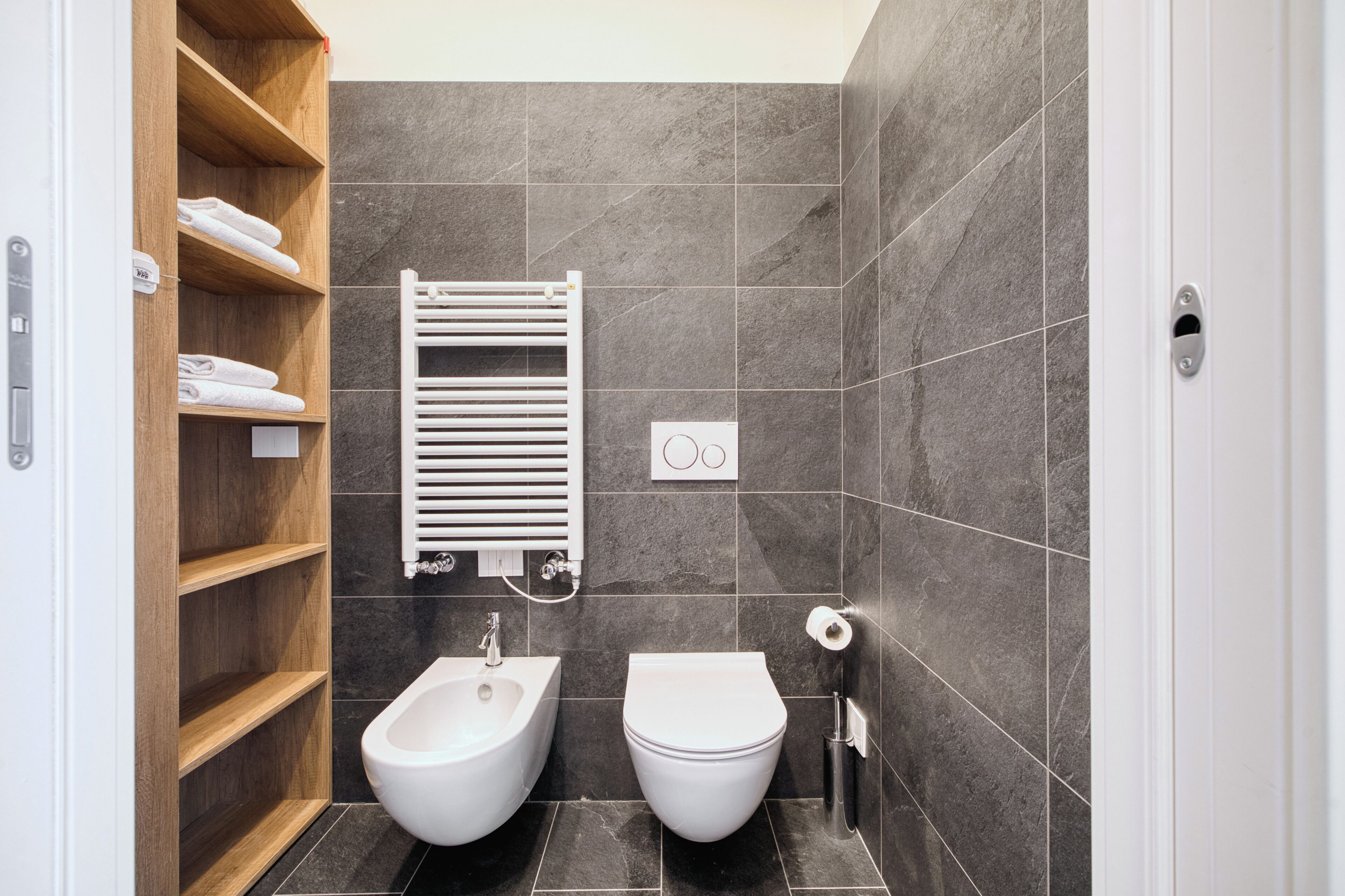 A modern bathroom with a bidet, toilet, and wooden shelves with towels.