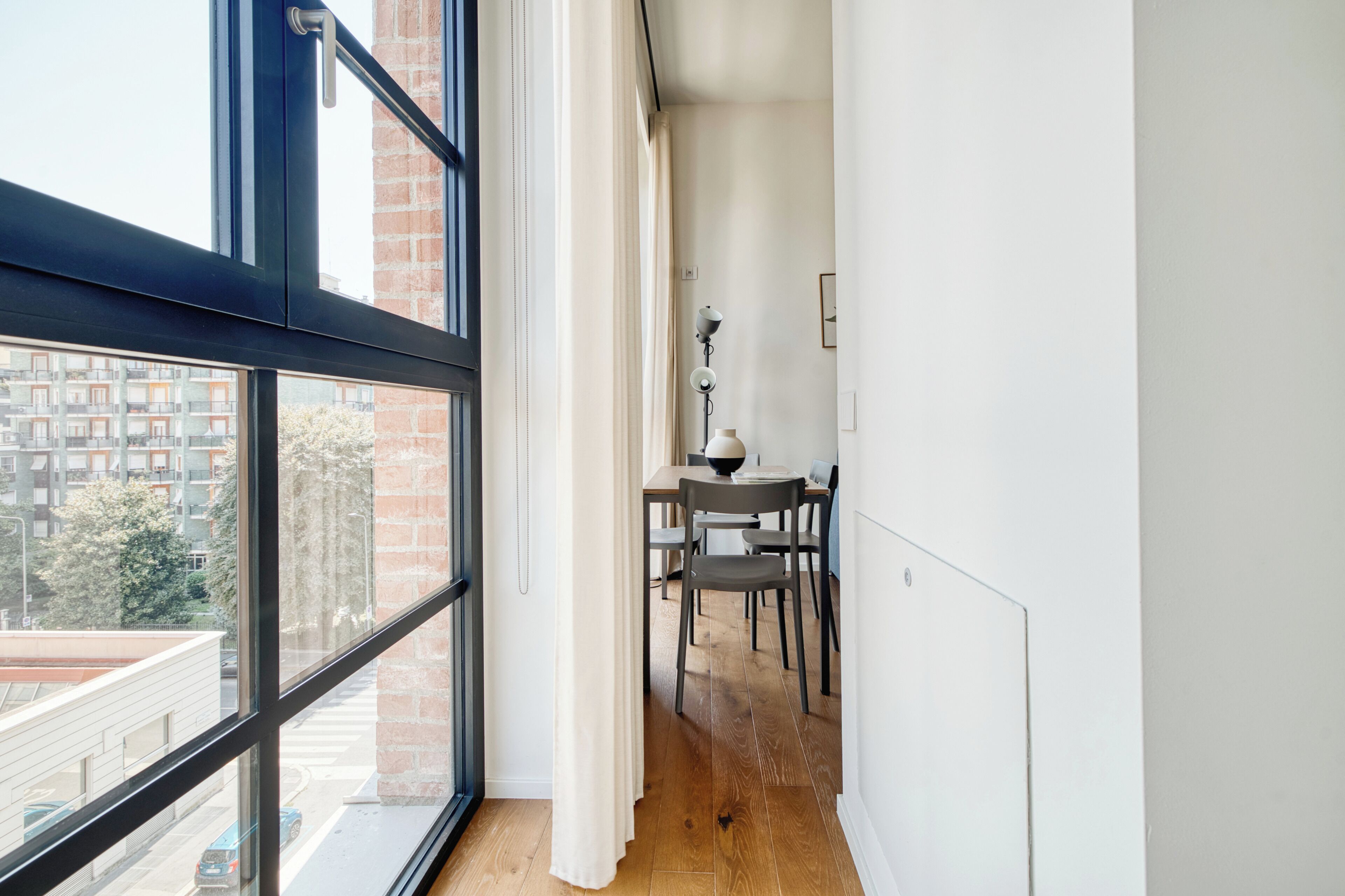 A narrow corridor with a dining area featuring a table and chairs, a lamp, and a view through large windows.
