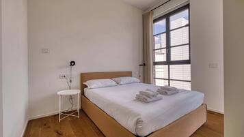 Une chambre à coucher avec un lit, une petite table de chevet blanche, une lampe et une grande fenêtre.