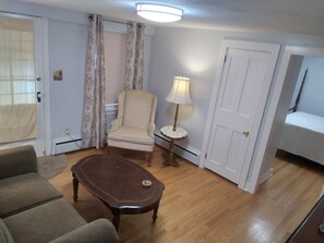 A room with a wooden floor, a beige armchair, a dark wooden coffee table, a small round table with a lamp, and a bed with a patterned bedspread. - Apartment for the traveling professional (Bristol)