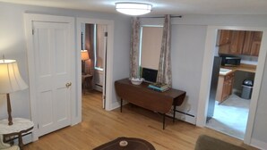 A room with a wooden floor, a table with a lamp, a chair, and a small table with books. There is a door leading to another room. - Apartment for the traveling professional (Bristol)