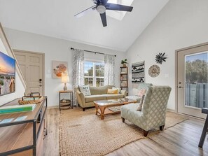 A living room with a ceiling fan, a sofa, a patterned armchair, a coffee table, and a large window with curtains. - Castaway Beach Bungalow Pets Welcome (Surfside Beach)