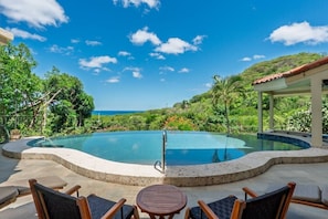 A pool with a curved edge, surrounded by chairs and a table, with a view of the ocean and hills in the background.