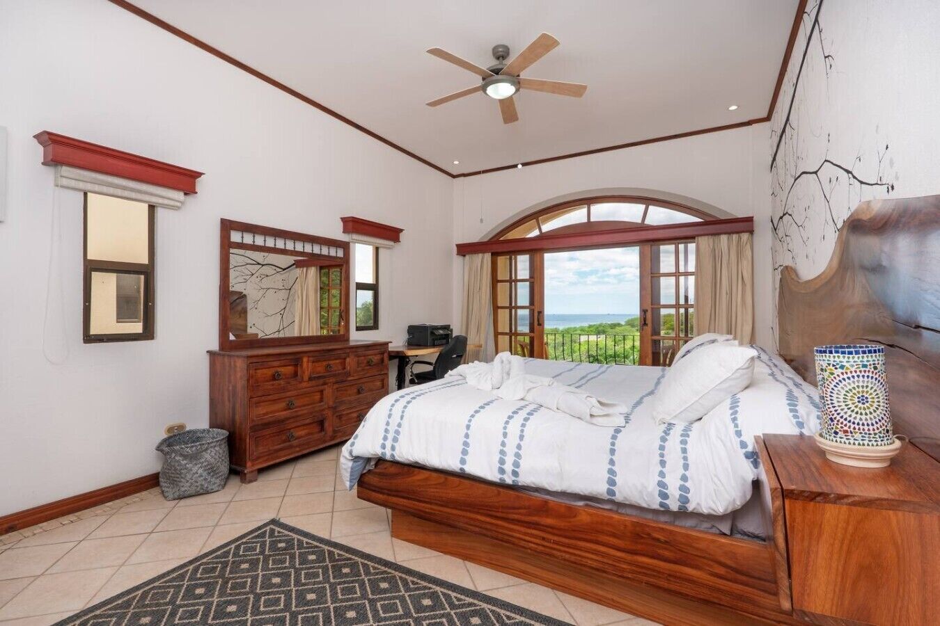 Ein Schlafzimmer mit einem großen Bett, einer Holzkommode, einem Schreibtisch und Blick ins Freie.