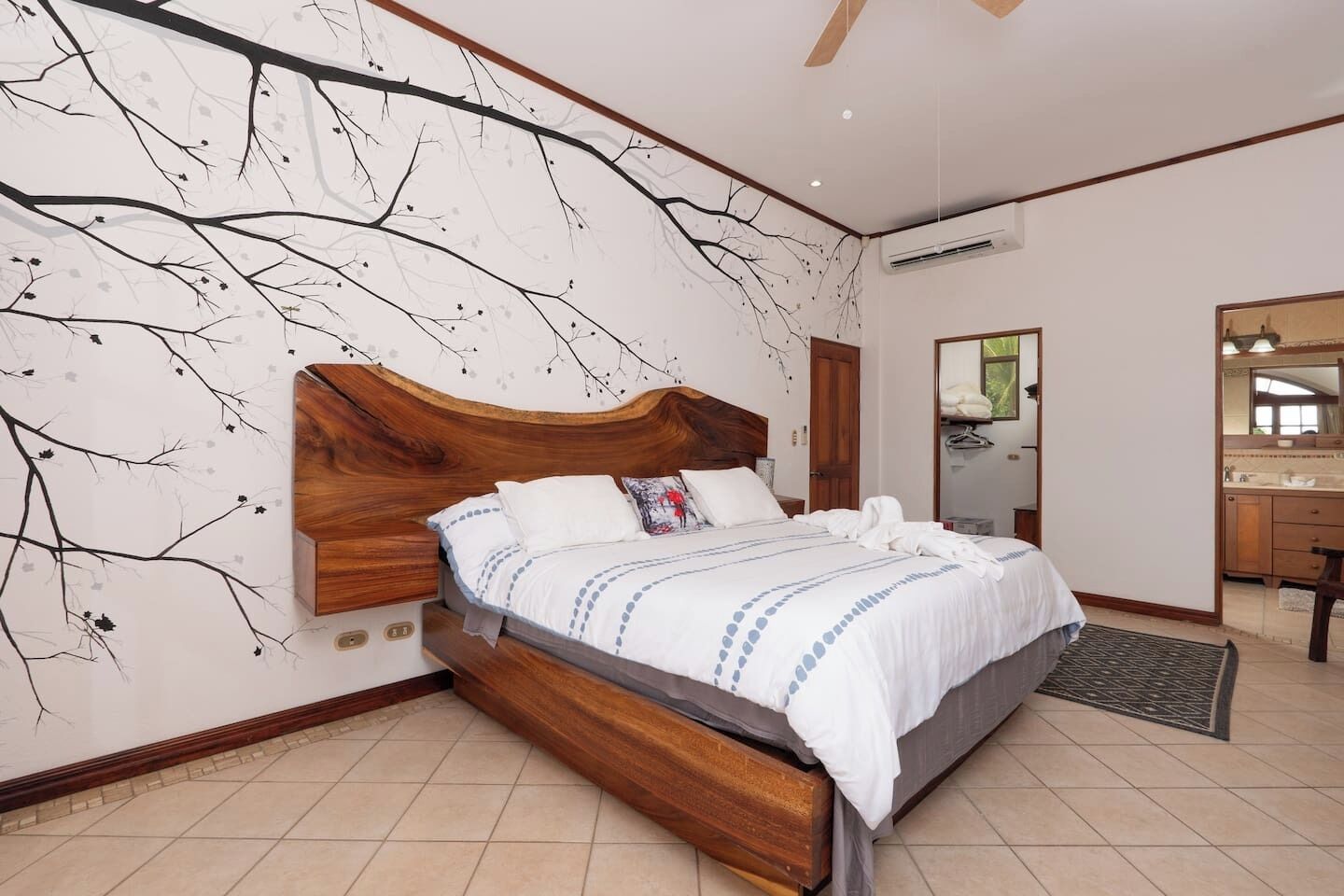 A bedroom with a large bed, a wooden headboard, a wall mural, a bathroom with a mirror, and a ceiling fan.