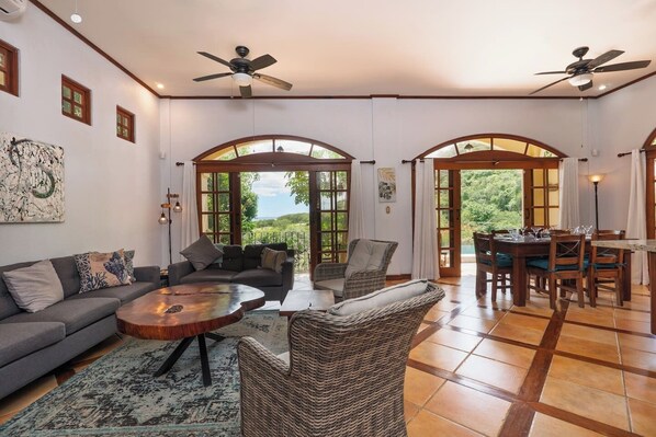 A spacious living area with a sofa, coffee table, and wicker chairs. There are arched doorways leading to an outdoor area.