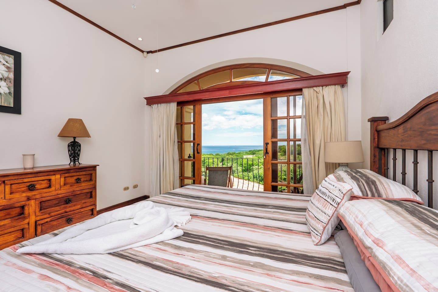 A bedroom with a large bed, a wooden dresser, and a view of the ocean through an open window.