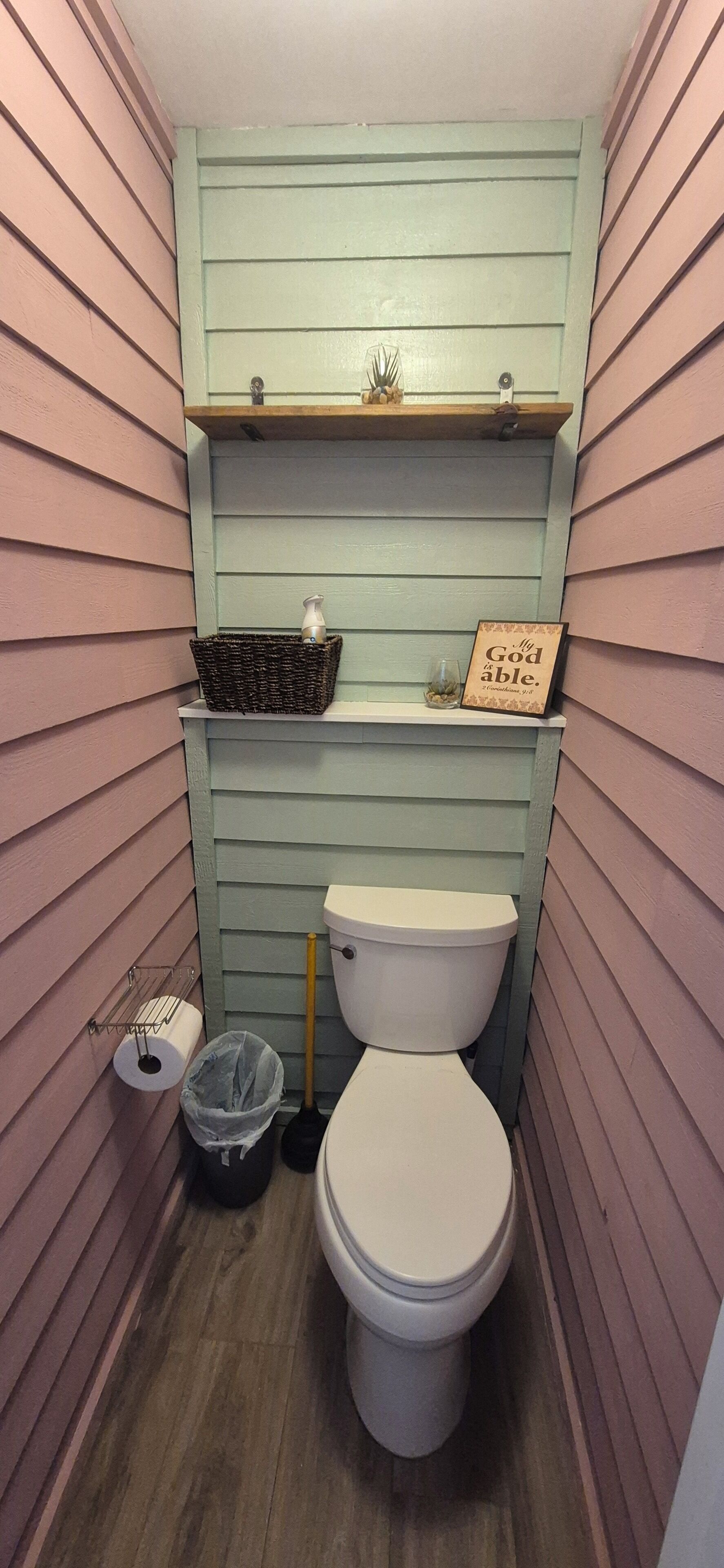 A small, narrow bathroom with a toilet, a shelf with decorative items, and a trash can.