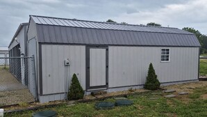 Een gelijkvloerse metalen schuur met een deur en een klein raam, omringd door gras en enkele potplanten.