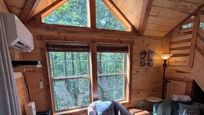 Ein gemütliches Blockhäuschen-Interieur mit Holzbalkendecke, zwei großen Fenstern, einer Couch, einem Sessel, einem kleinen Tisch und einer Lampe.