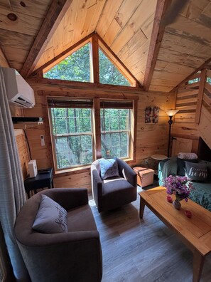 Ein gemütliches Blockhäuschen-Interieur mit Holzbalkendecke, zwei großen Fenstern, einer Couch, einem Sessel, einem kleinen Tisch und einer Lampe.