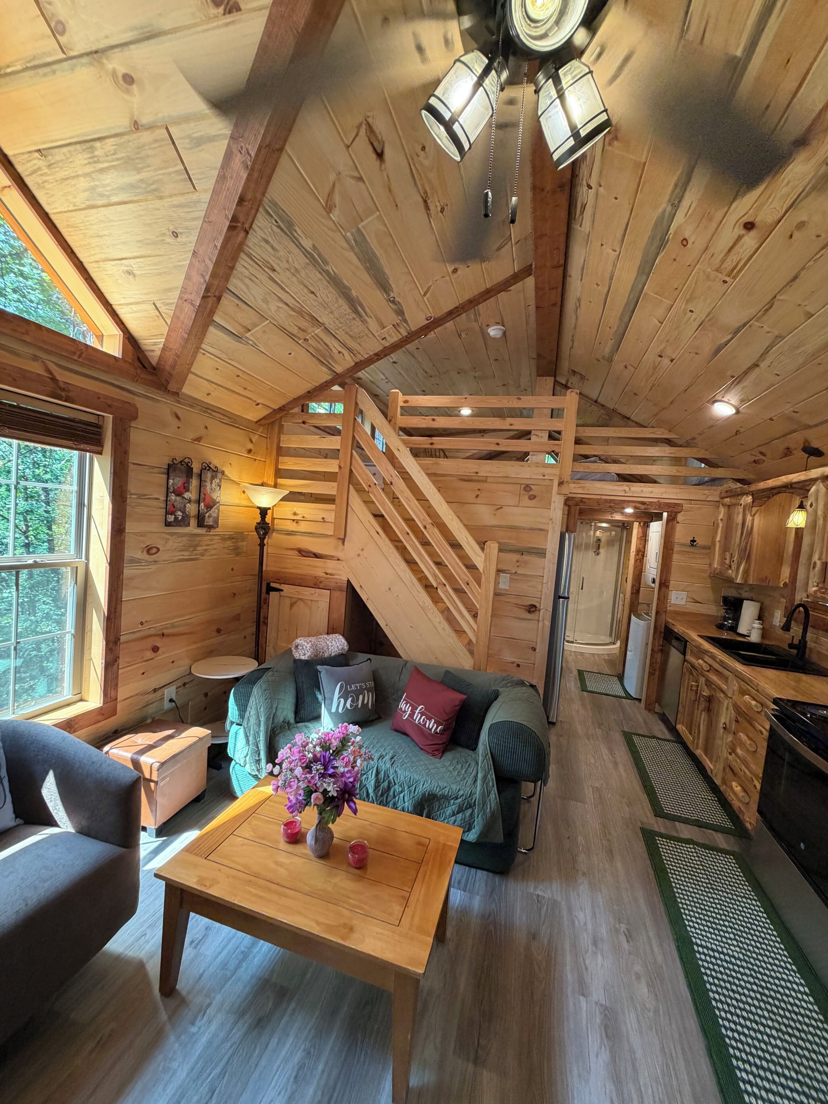 A wooden cabin interior with a staircase, a sofa, a coffee table with flowers, and a kitchen area.