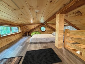 Ein gemütliches Schlafzimmer im Dachgeschoss mit schrägen Holzwänden, einem Einzelbett, einem Schaukelstuhl und einem Fenster mit Blick auf Grünflächen.