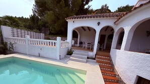 Una piscina con pared exterior blanca, un patio con mobiliario de exterior y una escalera de piedra que lleva a una entrada.