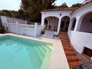 Ein Schwimmbad mit weißer Außenwand, eine Terrasse mit Gartenmöbeln und eine Steintreppe, die zu einem Eingang führt.