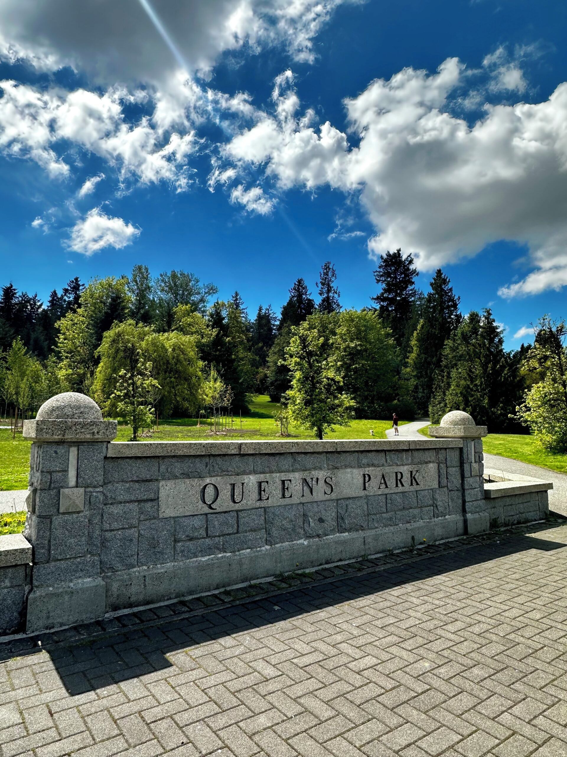 A stone wall with 'QUEEN'S PARK' engraved, surrounded by greenery and trees.