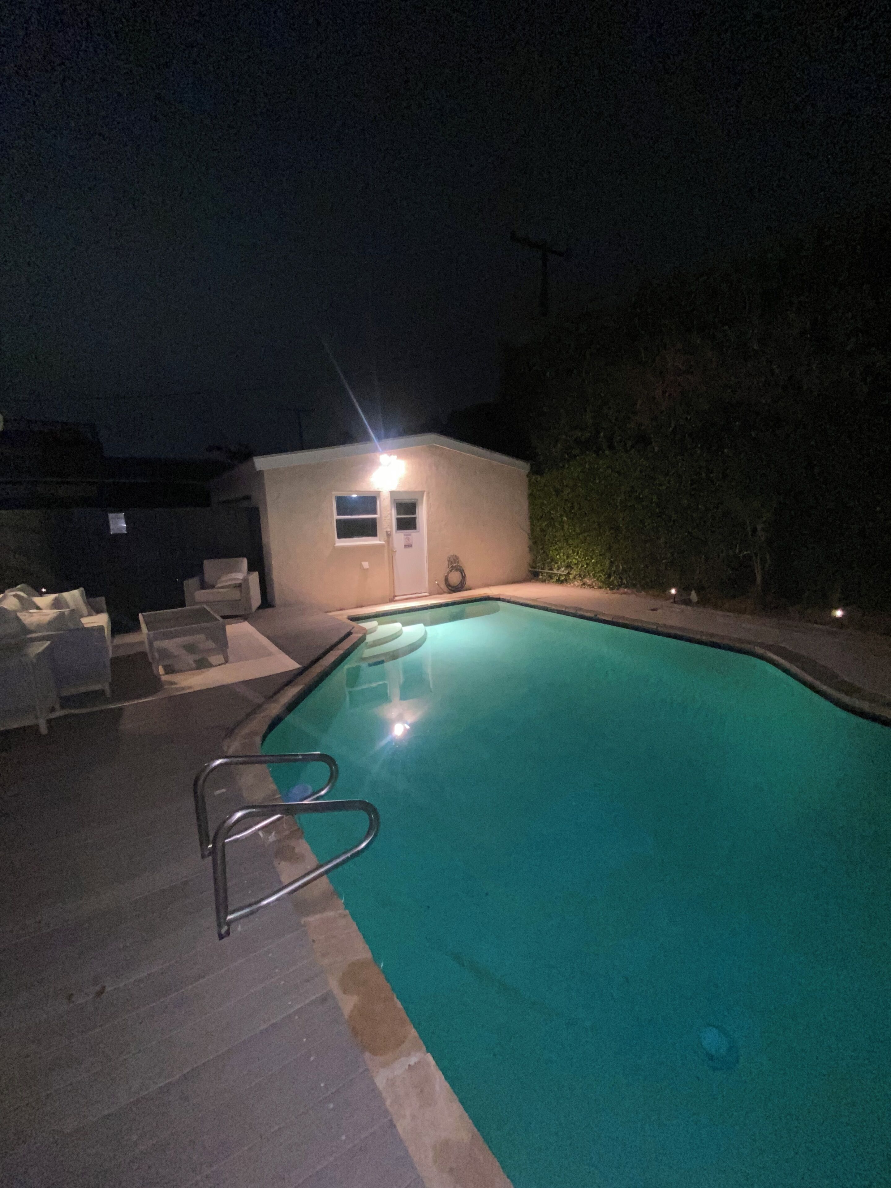 A swimming pool with a ladder, surrounded by lounge chairs and a small building with a door and windows.