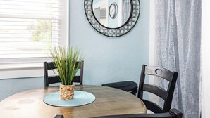 A round dining table with a woven centerpiece, two black chairs, a wall-mounted mirror, and a window with blinds. - Quiet Palm Loft Near Lake w/ BBQ & Patio (Lakeland)