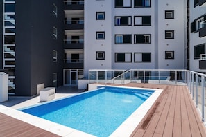 Ein Schwimmbecken mit Holzterrasse und ein modernes Gebäude mit Balkonen im Hintergrund.