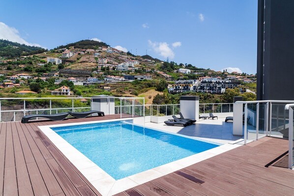 A swimming pool with lounge chairs on a wooden deck, overlooking a hillside residential area. - GuestReady - Family apartment with pool (Caniço)