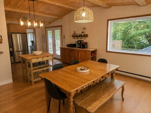 Una mesa de madera con sillas, una zona de cocina con refrigerador y una ventana con vista a árboles.