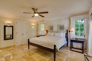 Une chambre à coucher comprenant un lit, un ventilateur de plafond, une table de chevet, une lampe, un miroir et une fenêtre avec des rideaux.
