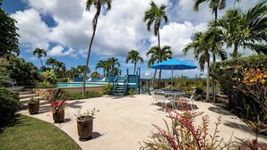 A pool area with lounge chairs, a blue umbrella, and palm trees. - Caribbean Blue Hideaway | 1BR Condo w/ Ocean View (Christiansted)