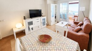 A modern living room with a flat-screen TV, a dining table with a patterned tablecloth, a sofa, and a balcony with a view. - Las Arenas T2 - Apartment with sea view (Isla Canela)