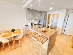 A modern kitchen with a central island, white cabinetry, and a dining area with wicker chairs. - Las Arenas T2 - Terrace with views (Ayamonte)