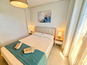 A neatly made bed with a blue blanket and a towel, two bedside lamps, a painting on the wall, and a window with curtains. - Las Arenas T2 - Terrace with views (Ayamonte)
