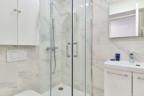A modern bathroom with a glass-enclosed shower, white marble tiles, a white sink, and a mirror above the sink. - Apartment in the Heart of the Marais (Paris)